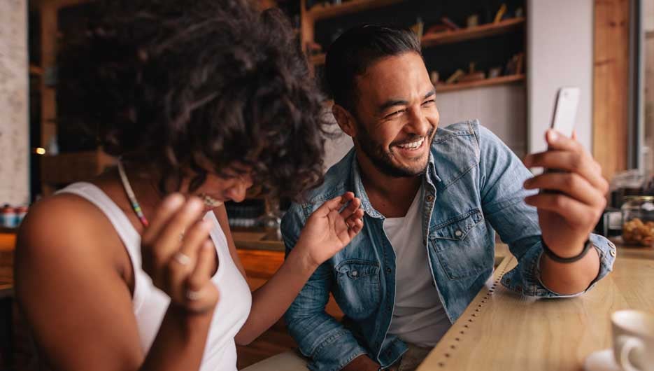 Smiling young couple sitting at coffee shop using mobile phone. Young man and woman at cafe taking selfie and laughing.